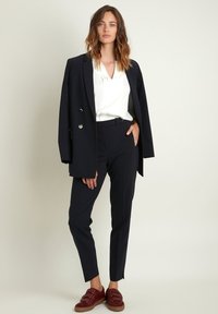 Blazer bleu marine à double boutons, blouse blanche, pantalon ajusté et chaussures slip-on rouges avec des bandes. Design épuré et minimaliste.