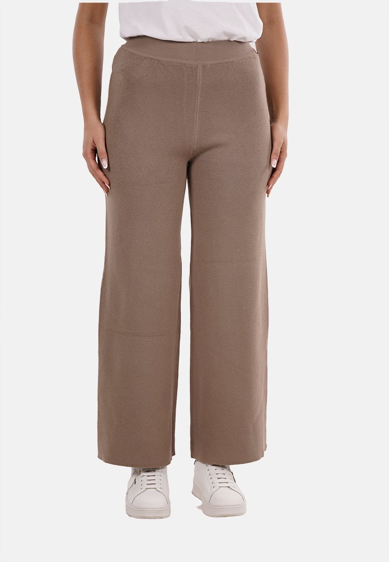 Pantaloni ampi e in maglia di colore marrone con una fascia in vita elasticizzata, caratterizzati da una texture liscia e un taglio alla caviglia, abbinati a sneakers bianche.