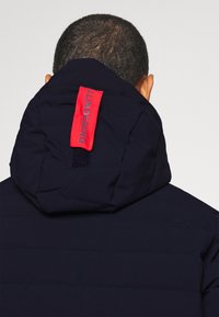 Marinblå puffjacka med en röd märkeslapp på huvan, med kviltad textur och en snygg, modern design.