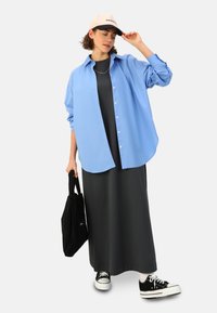 Chemise bleue à boutons portée par-dessus une robe maxi noire, accompagnée de baskets noires et d'une casquette beige. La mannequin tient un sac fourre-tout noir.