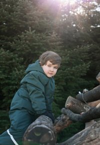 Copil într-o jachetă impermeabilă verde închis urcă pe bușteni de lemn. Buștenii texturați sunt aspri, cu o combinație de nuanțe de maro deschis și închis.