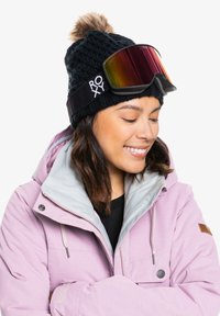 Musta neulepipo, jossa on keinoturkiskoriste, yhdistettynä sinisävyisiin hiihtolasiteillä, joissa on heijastava linssi. Vaaleanpunainen takki harmaalla vuorauksella.