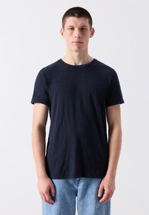 Mladý muž stojaci, oblečený v jednoduchom čiernom tričku s krátkym rukávom a svetlomodrých rifliach, na jednoduchom bielom pozadí.