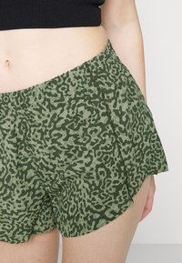 Calções verdes estampados com um cós elástico, apresentando um discreto padrão de leopardo em várias tonalidades de verde e um detalhe na costura lateral.
