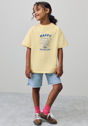 Geel T-shirt met blauwe grafische print, gecombineerd met lichte denim shorts. Felroze sokken en schoenen met luipaardprint maken de outfit compleet.