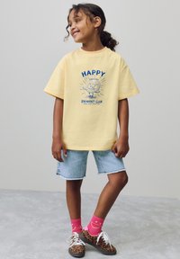 T-shirt gialla con stampa grafica blu, abbinata a pantaloncini di denim chiaro. Calzini rosa acceso e scarpe con stampa leopardata completano l'outfit.