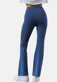 Dopasowane niebieskie legginsy w stylu dzwonów wykonane z elastycznego materiału, z elastycznym pasem w talii i gładką fakturą, w połączeniu z białymi sneakersami.