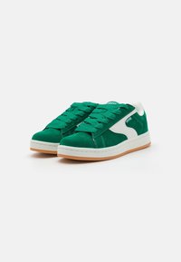 Baskets en daim vert avec des lacets plats verts, des accents blancs, une surface texturée et une semelle extérieure en caoutchouc naturel. Design décontracté, silhouette basse.