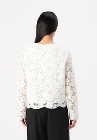Top bianco in pizzo a maniche lunghe con bordo ondulato e motivi floreali. Il tessuto è semitrasparente e sovrapposto a un indumento più scuro.