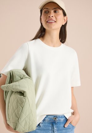 Jeune femme portant une casquette beige et un t-shirt blanc tient une veste matelassée vert clair sur le bras, debout devant un fond beige.