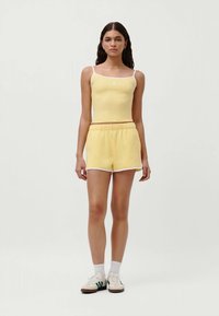 Ung kvinne iført en gul tank topp og matchende shorts, stående strak med hvite sokker og joggesko mot en ensfarget bakgrunn.