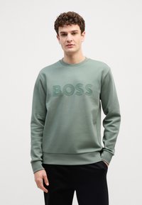 Felpa verde con collo a giro, caratterizzata da un logo "BOSS" in rilievo sul davanti. Realizzata in tessuto morbido e testurizzato, presenta polsini e bordo a costine.