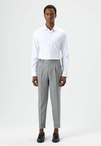 Witte blouse met lange mouwen gecombineerd met grijze, op maat gemaakte broek met een broekriemlus. De broek heeft voorplooien en opgerolde pijpen. Zwarte schoenen.