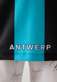 Fekete és türkiz sportpóló, az alján vastag fehér betűkkel a "ANTWERP" felirattal, sima, szőtt textúrával.