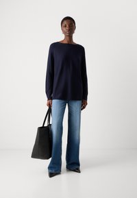 Marineblå langermet topp, avslappet passform; kombinert med lys blå vide jeans. En svart tote-bag holdes i høyre hånd, med en glatt tekstur.