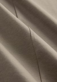 Smeđa tkanina glatke teksture, sa dijagonalnim naborima i suptilnim detaljima šavova, prikazuje meki, spušteni izgled.