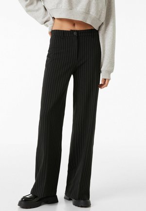 Zwarte pantalon met streepjes en een rechte pijp, voorzien van een knoopsluiting en zijzakken, gecombineerd met stevige zwarte schoenen.