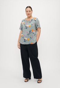 Blommig blus i ljust grönt tyg med korta ärmar; har rosa, orange och blå blommönster. Kombineras med lösa svarta byxor.