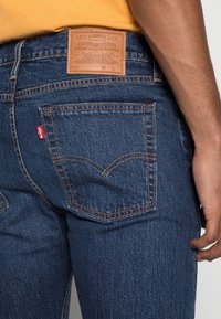 Vista trasera de una persona que lleva puestos unos pantalones vaqueros Levi's de un azul oscuro, con una etiqueta de marca de cuero marrón y una etiqueta roja de Levi's en el bolsillo, de pie frente a un fondo blanco.
