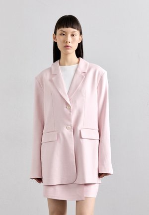 Blazer oversized rosa claro com dois bolsos frontais, fecho de botão único e textura suave, combinado com uma saia mini a condizer.