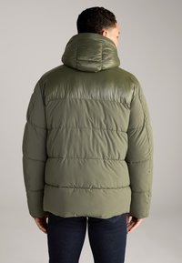 Olivenfarbene Steppjacke mit glänzender Oberfläche, ausgestattet mit Kapuze, gestepptem Design und elastischen Bündchen. Dunkelblaue Jeans sind am unteren Ende sichtbar.