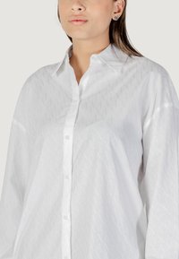 Camicia bianca a maniche lunghe con un sottile motivo geometrico. Presenta un colletto polo e bottoni frontali. Tessuto morbido, vestibilità comoda.