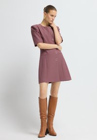 Vestido de ciruela de manga corta con detalles de botones y bolsillos laterales. Combinado con botas de cuero marrón hasta la rodilla que tienen una cremallera lateral.