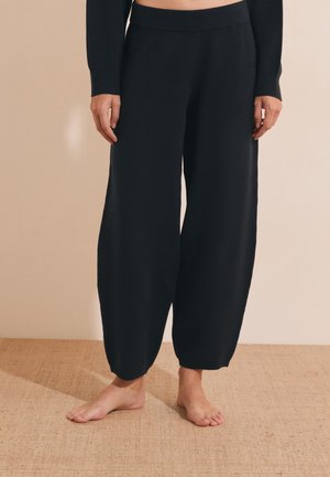 Persoon draagt zwarte wijde gebreide broek en een zwart cropped top, staat blootsvoets op een gestructureerde beige vloer tegen een effen muur.