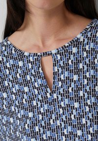 Donna che indossa una blusa a fantasia blu, nera e bianca con una piccola apertura a chiave davanti.