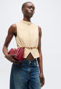 Γυναίκα με αμάνικη μπεζ μπλούζα με κοψίματα μπροστά και χρυσή ζώνη, κρατά κόκκινο δερμάτινο clutch, ντυμένη με μπλε τζιν πάνω σε λευκό φόντο.