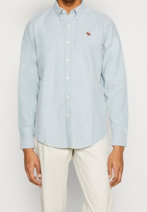 Mężczyzna w jasnoniebieskiej koszuli na guziki z haftowanym logo łosia na piersi i w jasnobeżowych spodniach, stojący na jednolitym tle.