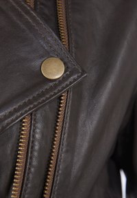 Bruine leren jas met messing hardware, met een diagonale rits en drukknopdetail. Gladde textuur met zichtbare stiksels.