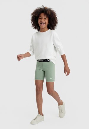 Barn med krøllet hår smiler, mens det går, iført en hvid sweatshirt, lysegrønne Nike-shorts og hvide sneakers på en ensfarvet baggrund.