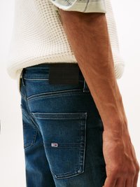 Primer plano de una persona que lleva vaqueros de mezclilla azules con un bolsillo trasero rectangular, un pequeño logotipo y la manga de un suéter de punto color crema visible.