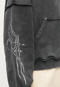 Camisola com capuz cinzenta escura com tecido texturizado, bolso lateral e destaque em design gráfico branco na manga. Detalhe de bainha com borda crua.