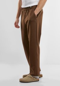Brown, lightweight fabric trousers with an elastic waistband, side pockets, and a relaxed fit. Worn with beige clogs and white socks.
