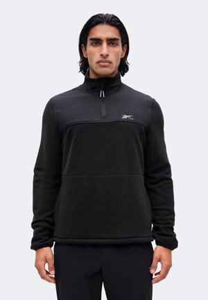 Mand med sort hår iført en sort langærmet pullover med en halv lynlås og Reebok-logo på brystet, stående mod en lys baggrund.