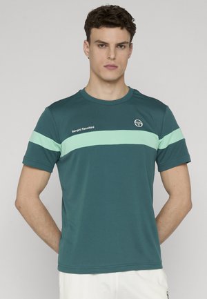 Ung mand med kort, krøllet hår iført en teal Sergio Tacchini t-shirt med en lys grøn vandret stribe og hvide shorts.