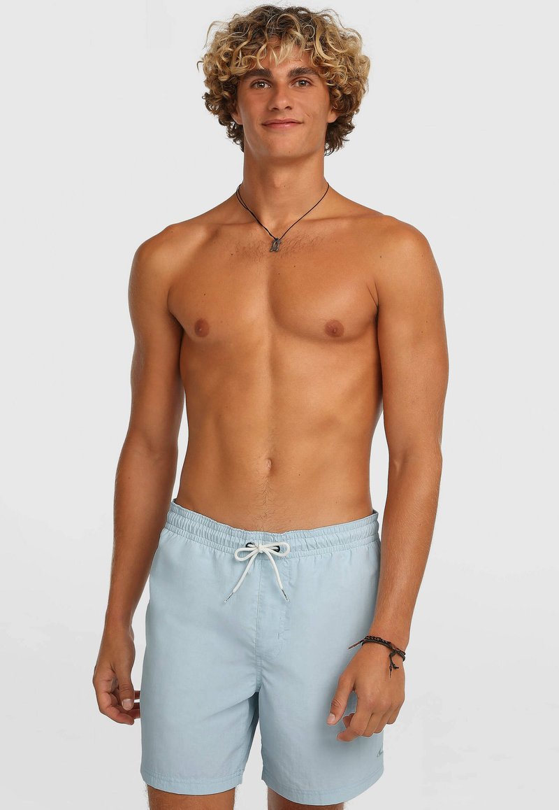 Giovane uomo con capelli ricci che indossa pantaloncini da bagno azzurri e una collana, in piedi davanti a uno sfondo semplice.