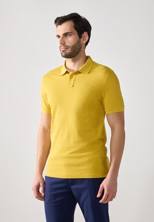Camiseta polo amarilla de manga corta con cuello clásico y una única botonera. Textura suave, tela ligera, combinada con pantalones azules.