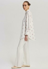 Camicia bianca a maniche lunghe con motivi floreali rossi, colletto abbottonato e vestibilità rilassata, abbinata a pantaloni bianchi a palazzo e ballerine beige.