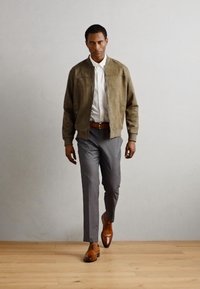 Giacca bomber in suede marrone sopra una camicia bianca con colletto, pantaloni grigi su misura e scarpe in pelle marrone. Pavimento in legno, sfondo neutro.