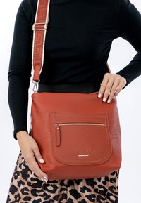 Rode crossbodytas van getextureerd leer met een voorvak met ritssluiting en een verstelbare band met patroon. Merkennaam zichtbaar aan de voorkant.