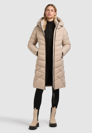 Beige gewatteerde jas met capuchon, diamantpatroon, voorrits en zijzakken, gedragen over een zwarte outfit met beige laarzen.