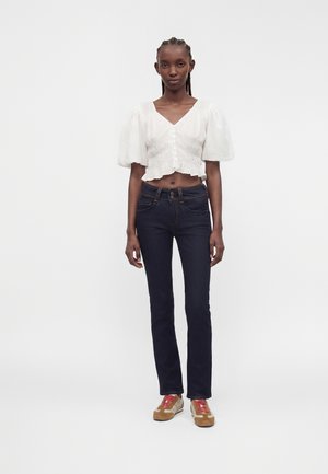Femme portant un chemisier court blanc à manches bouffantes, un jean bleu foncé et des baskets marron avec des lacets rouges, debout devant un fond uni.