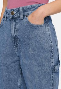 Pessoa a usar jeans de ganga azul de cintura alta com a mão no bolso da frente e uma blusa rosa por dentro.