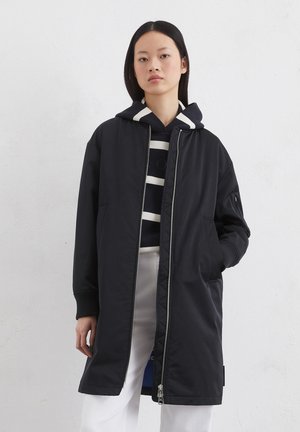 Black longline jacket with a zip closure, ribbed cuffs, and side pockets. Worn over a striped navy and cream hoodie. Smooth texture.