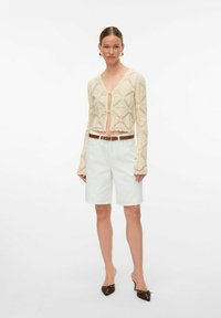 Licht beige cropped cardigan met textuurpatronen, voorzien van lange flared mouwen en een sluiting met frontstrik. Gekleed met witte op maat gemaakte shorts.