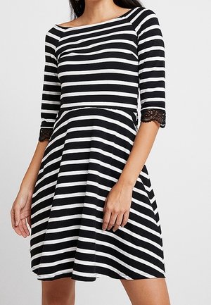 Femme portant une robe à rayures horizontales noires et blanches avec des manches mi-longues et un galon en dentelle aux poignets.