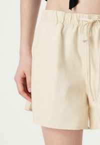 Lys beige shorts med elastisk midjebånd og snor. Stoffet ser glatt ut med en lett rynket tekstur.